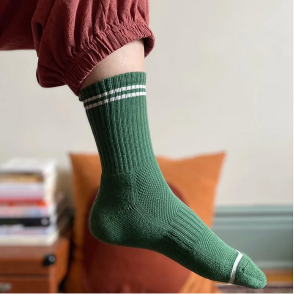 A person wearing a ribbed green crew sock with white stripes near the top and a white toe cap, shown in a styled indoor setting with a rust-colored pants leg visible.