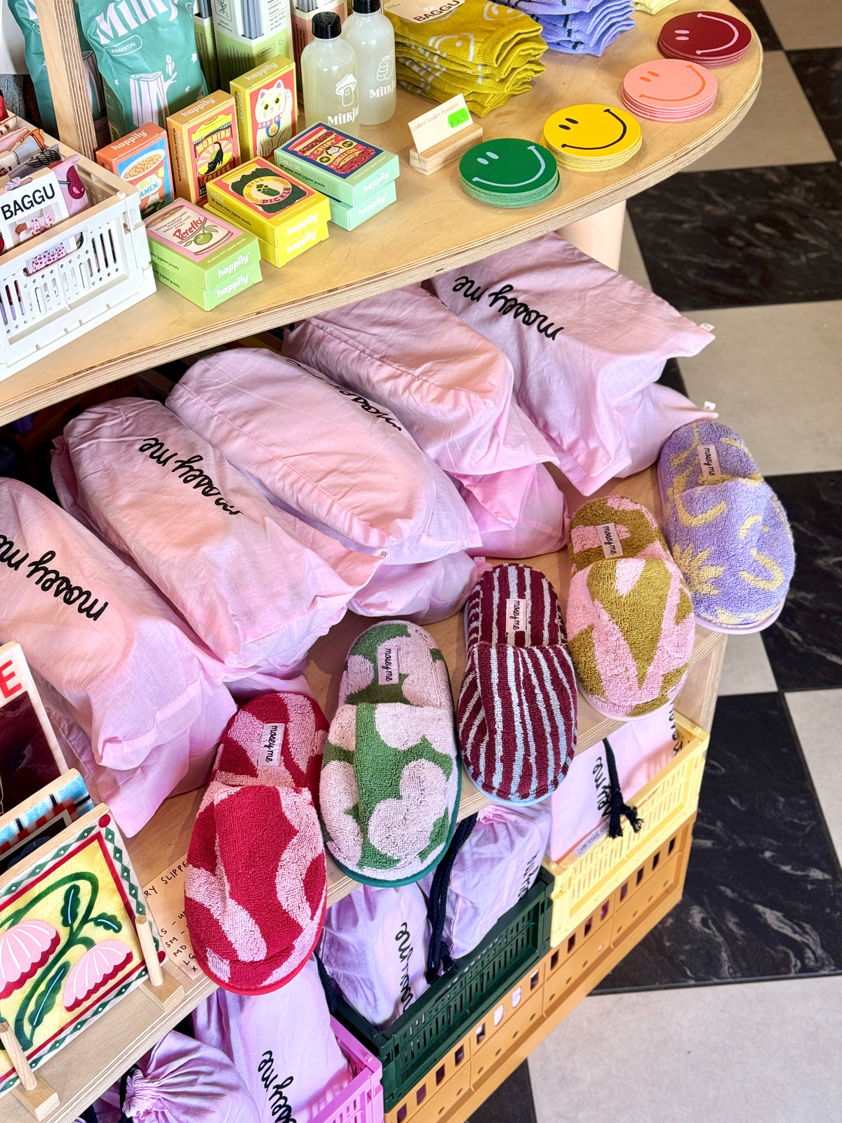 A retail display shelf filled with multiple pairs of colorful knit slippers and matching soft pouches, with small boxed items on the upper curved shelf. The slippers come in several colors including pink, green, red, purple, and striped patterns, and the display is photographed in a store setting with a black-and-white checkered floor visible.