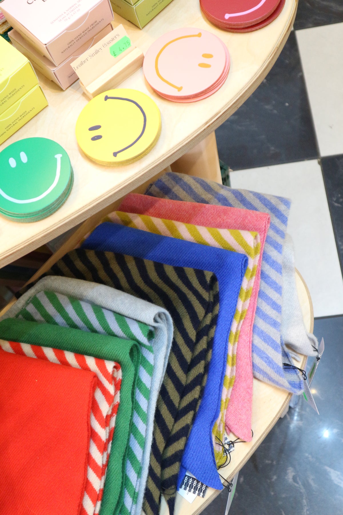 A close-up view of folded colourful woollen scarves arranged on a display table, with smiley-face shaped items on a wooden shelf beside them.
