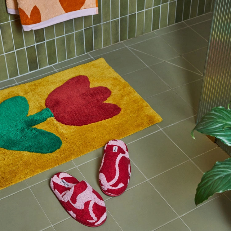 Yellow bath mat with red and green floral design on a tiled bathroom floor, accompanied by red slippers.